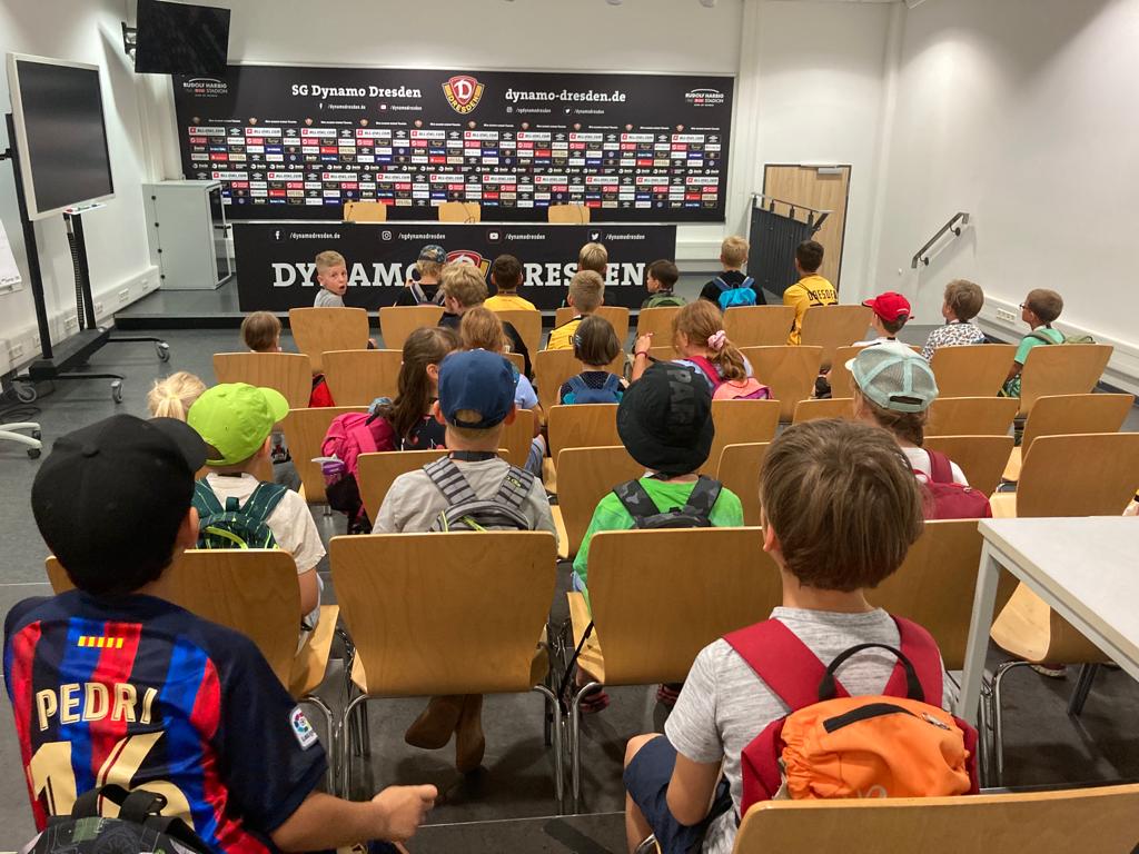 Kinder sitzen in einem Raum vor einem Podium, welches vor einer schwarzen Wand mit Sponsorenlogos steht, am Podium hängt ein schwarzes Banner mit der Aufschrift Dynamo Dresden
